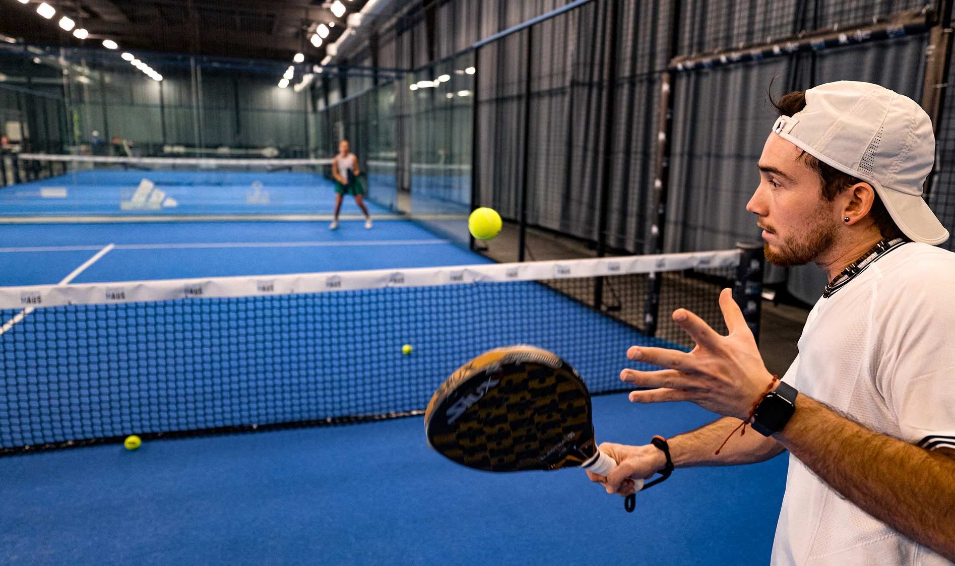 Person playing paddle tennis on an indoor court with another player in the background.